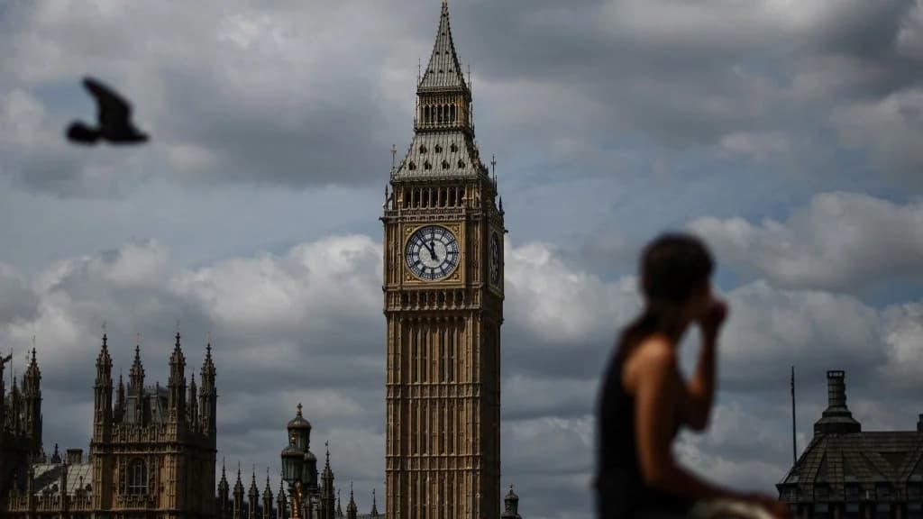A woman sits on the south side of the River Thames with a view of Big Ben and the Palace of Westminster, London, 19 July 2026 (AFP)
